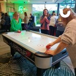 Air Hockey Table being used at an office party by 2 competitive women with another woman looking on in delight
