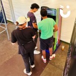 Arcade Machine in a marquee at a daytime event with 2 friends playing and a person watching on