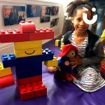 A young girl sitting next to here Lego Paddington Bear whilst holding a Teddy version of Paddington