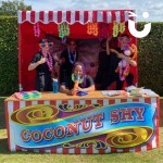 Coconut Shy Stall Hire set up on a sunny day with 5 fun experts laughing and posing with prizes