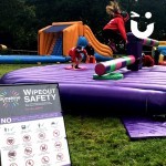 Guests of an outdoor family fun day take on the challenge of the wipeout. A young girl jumps over the sweeper arm as the next competitor steadies them self ready.