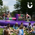 Guests of a family fun day enjoy the sunshine while behind them, others take on the challenge of the wipeout inflatable hire from Sunshine Events