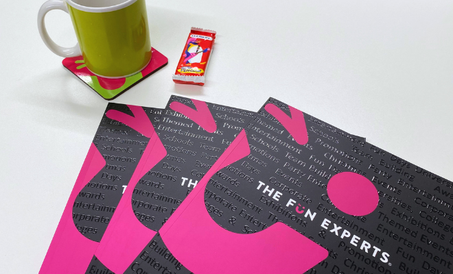 Three Event Entertainment Guides fanned out on a table with a green mug on a Fun Experts coaster and a penguin biscuit beside it